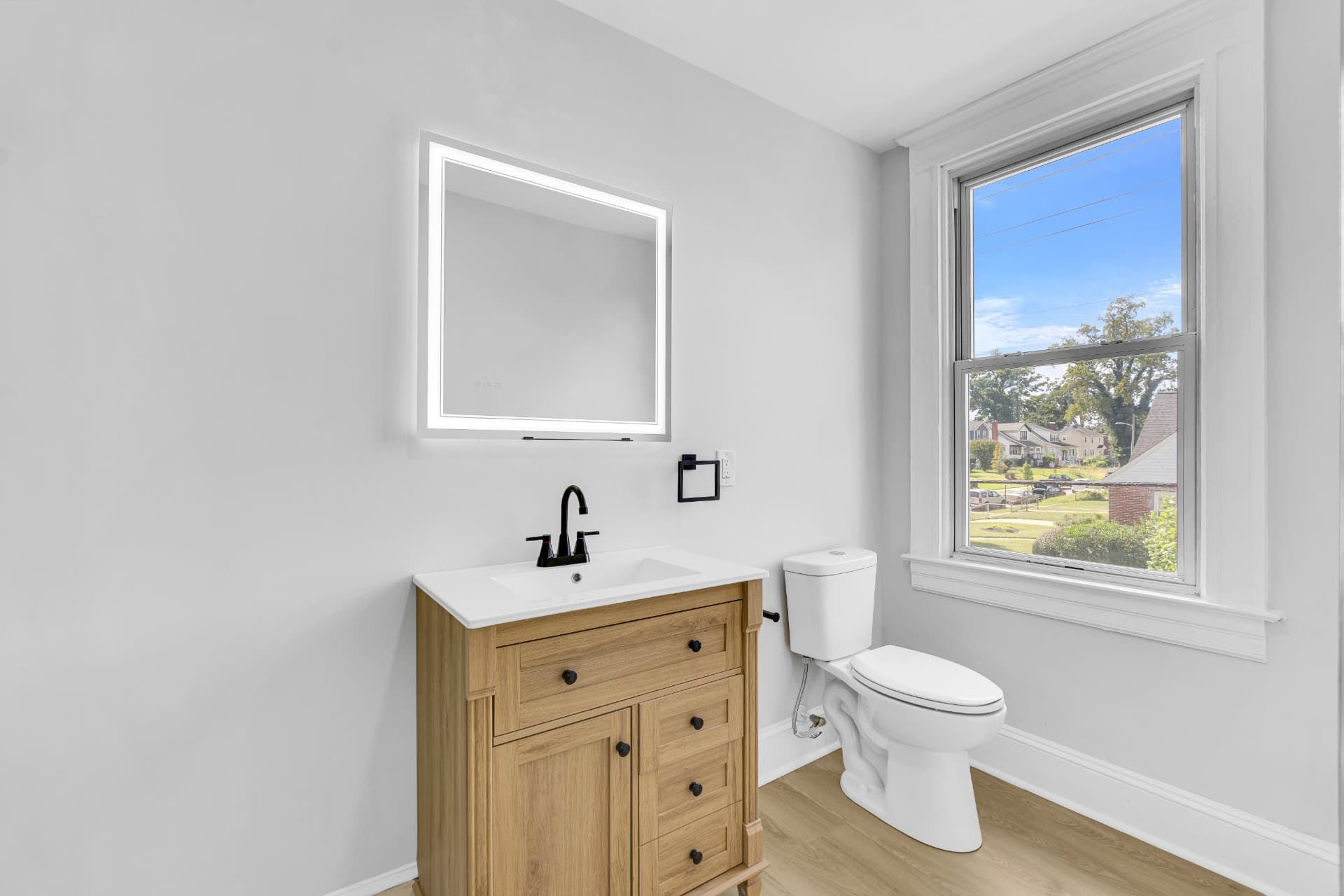 Powder room with a wood vanity, backlit LED mirror and a sunny neighborhood window view